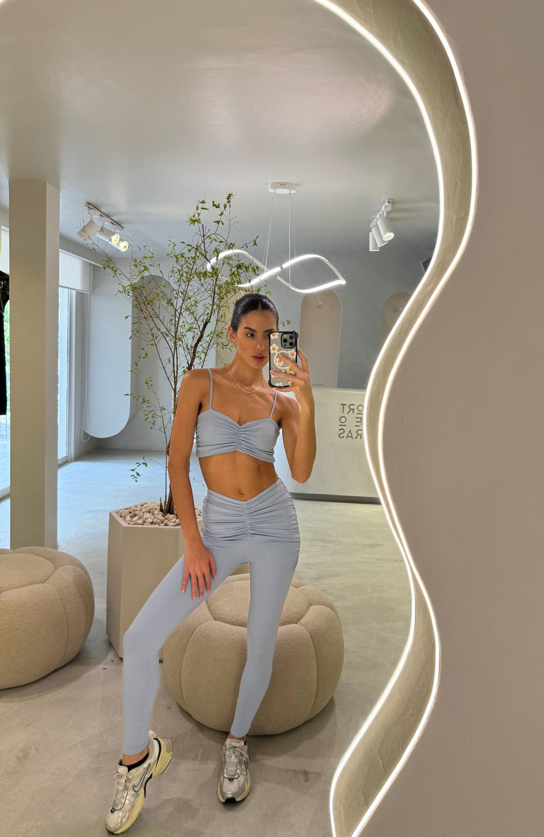 A person wearing light blue leggings and a sports bra, sitting in a modern room with a plant and a mirror in the background.