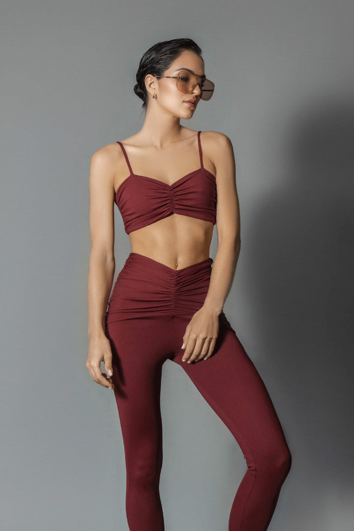 A female model wearing a burgundy sports bra and matching leggings, standing against a grey background.