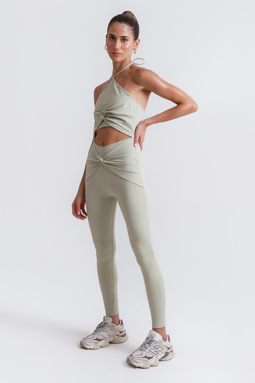 A model wearing a light green unitard with a halter neck and cut-out at the waist, paired with matching leggings and sneakers.