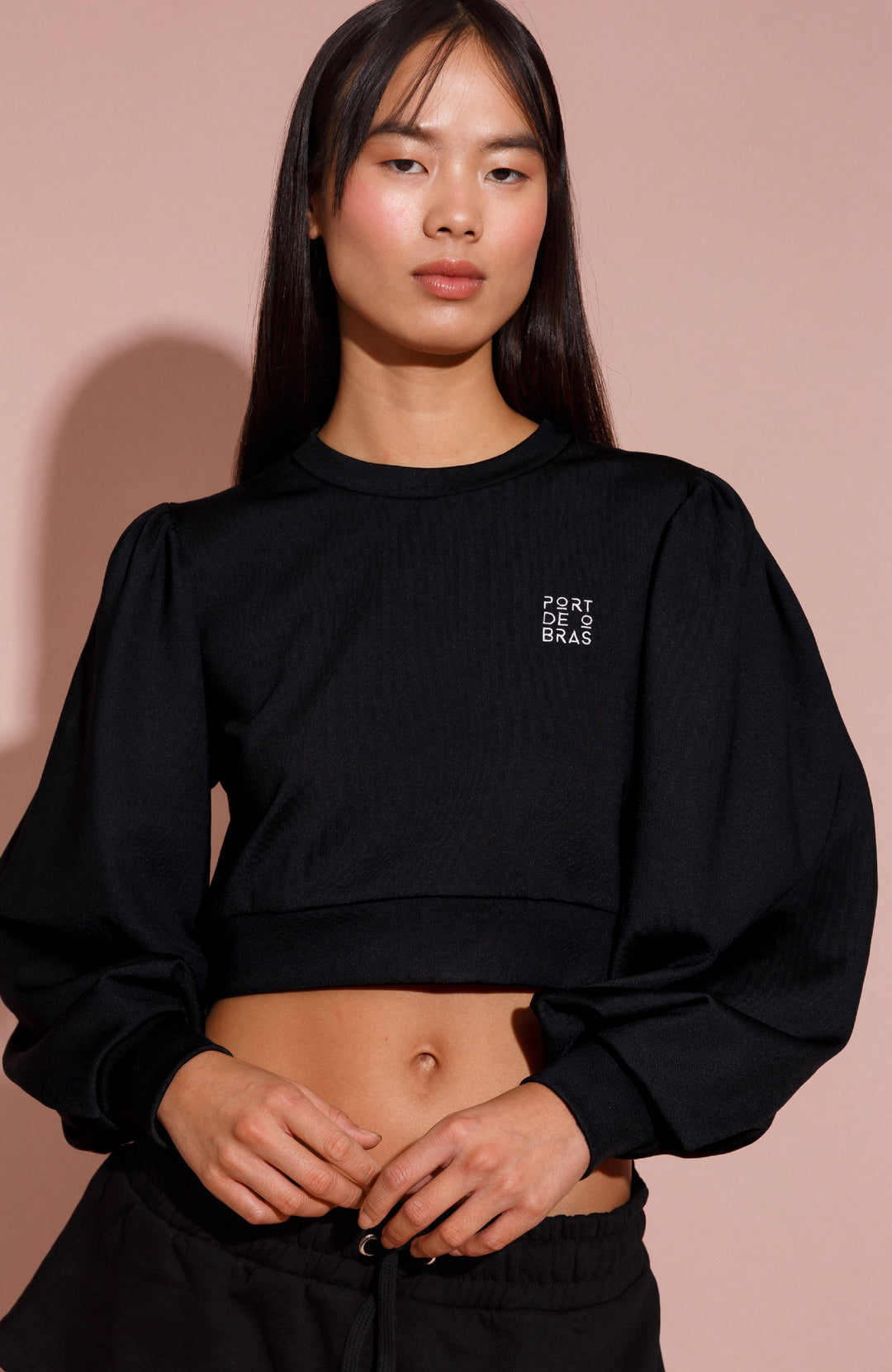 A woman wearing a black cropped sweater with long sleeves, featuring a small logo on the upper left chest area.