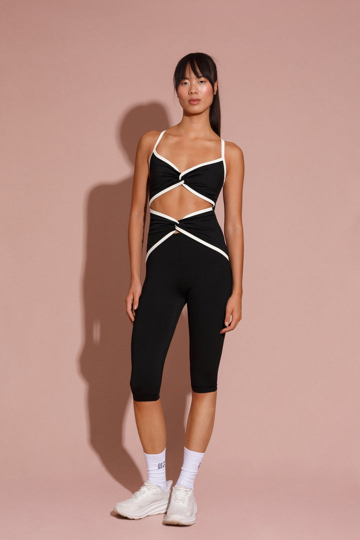 A model wearing a black ribbed capri suit with white cutouts at the chest and waist, paired with white sneakers and socks.