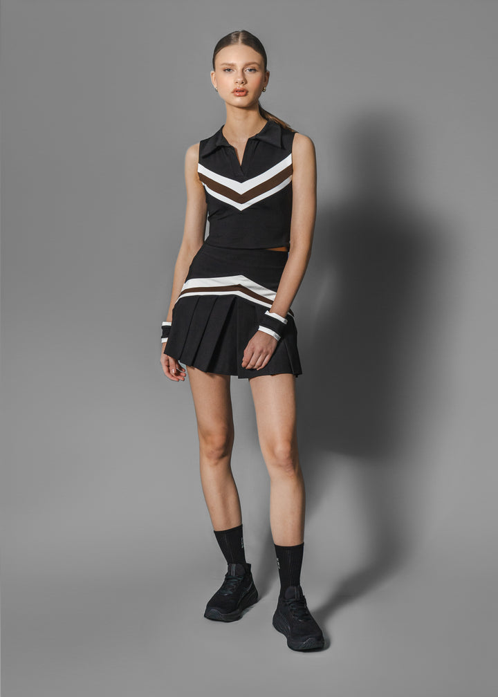 A female model wearing a sleeveless black top with white and chocolate brown geometric patterns, paired with a matching tennis skirt and socks.