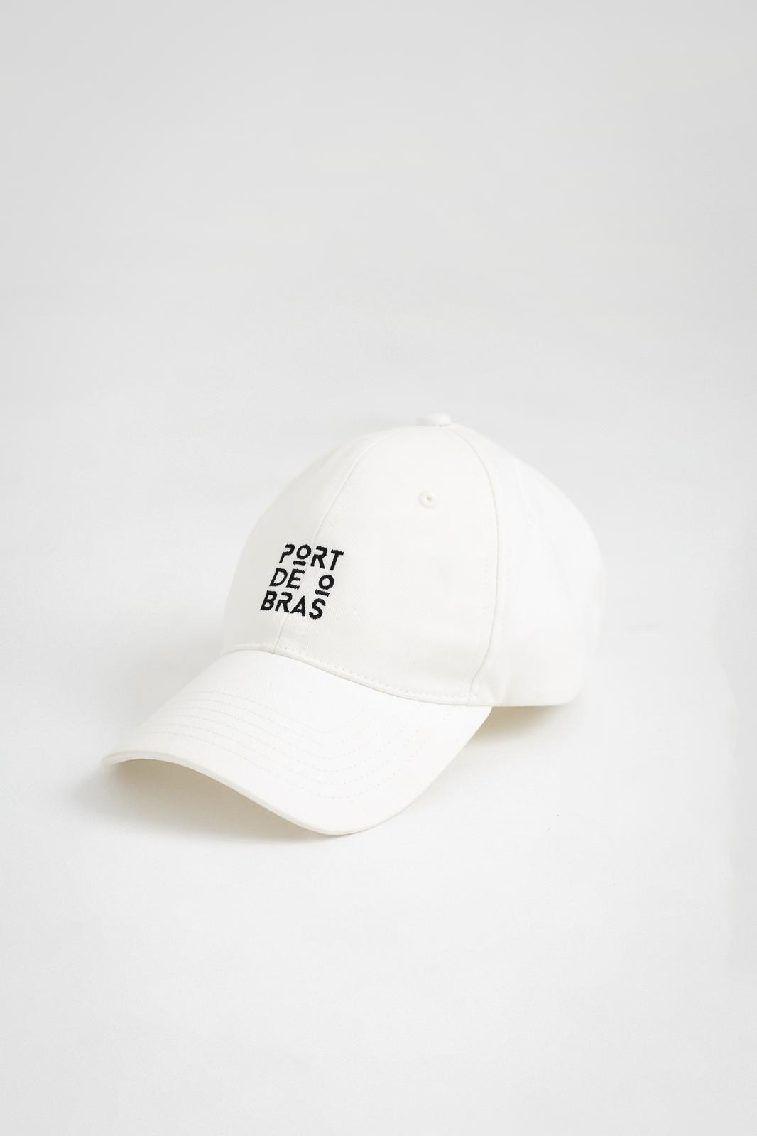 A white baseball cap with 'PORT DE BRAS' written in black text on the front.