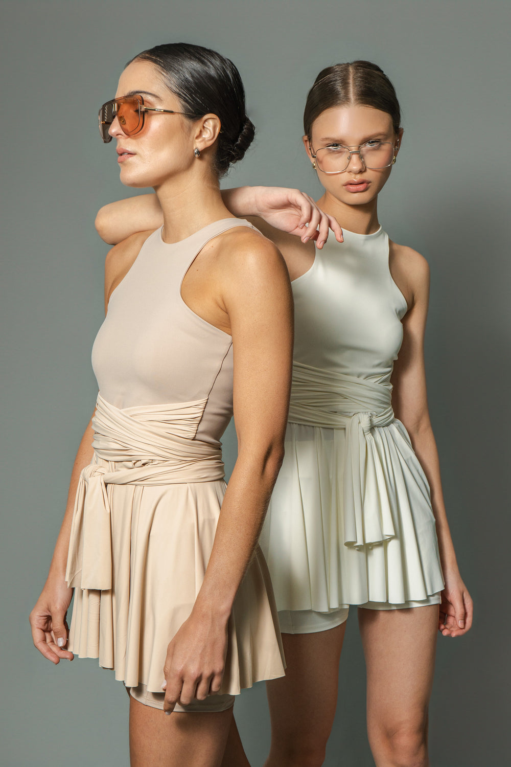 Two models wearing sleeveless dresses, one in beige and the other in white, both with sunglasses and posing against a neutral background.