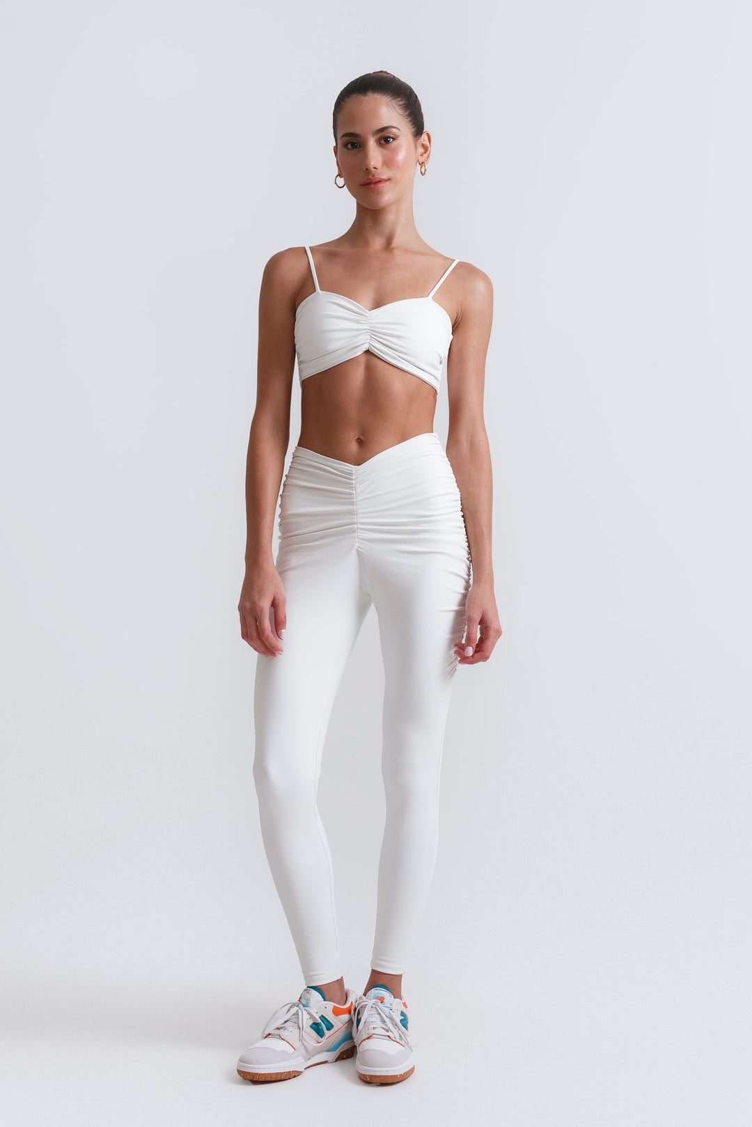 A woman is standing, wearing white Ariadne leggings with a matching white top. She has accessorized with earrings and is standing against a white background.