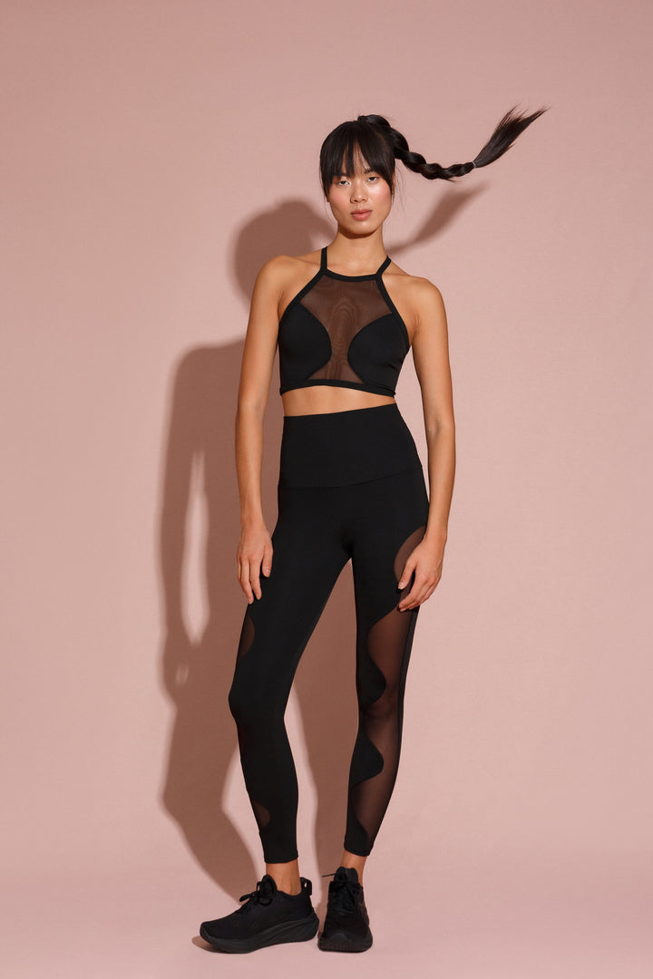 A female model wearing a black high-waisted leggings and a matching sports bra with mesh detailing. The outfit has a high neck and sleeveless design with a ballet-inspired silhouette.