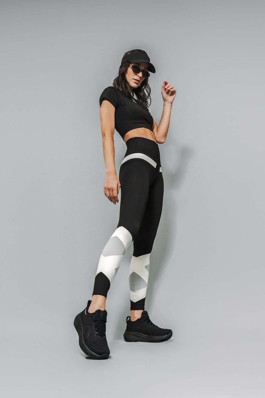 A model wearing high-waisted black leggings with symmetrical white and gray geometric patterns on the lower legs, paired with a black crop top and black sneakers.