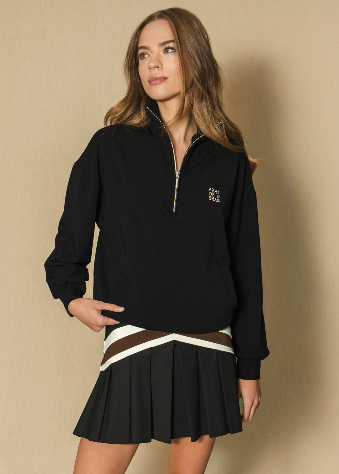 Woman wearing a black quarter-zip sweatshirt and pleated skirt against a beige background