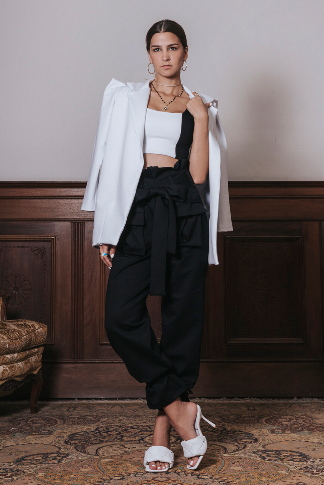 A female model wearing black cargo trouser pants with a white blazer and white heels. Accessories include a necklace and earrings.